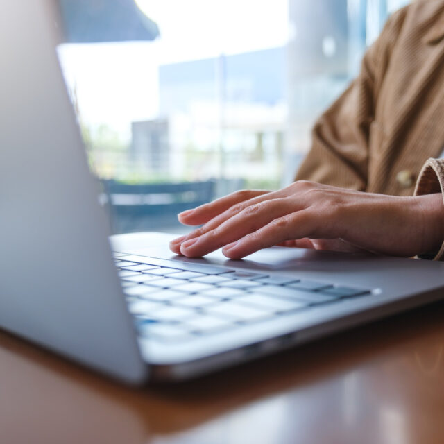 A person wearing a brown jacket types on a silver laptop on a wooden table in a bright room.