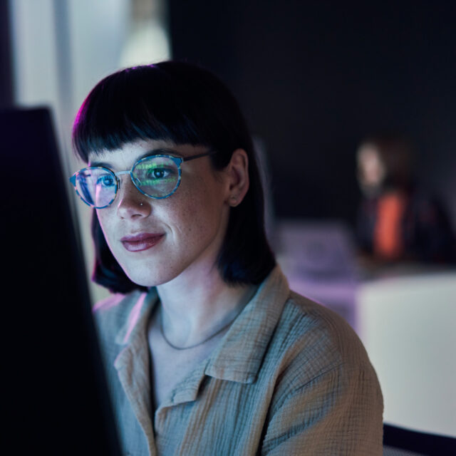 A person with short black hair and glasses is sitting at a desk and looking at a computer screen in a dimly lit room.
