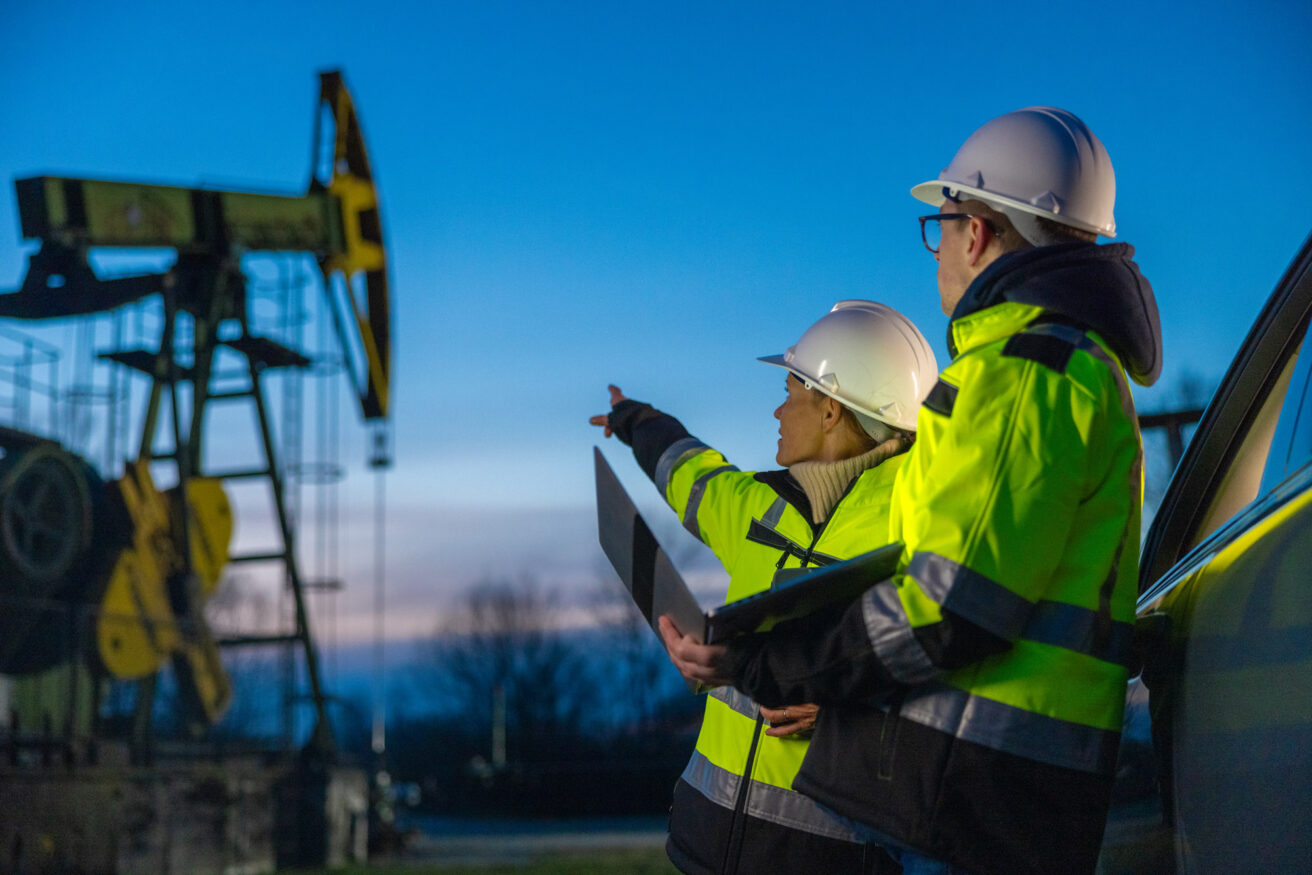 Two workers in high-visibility jackets and hard hats point towards oil extraction machinery at dusk, holding a laptop.