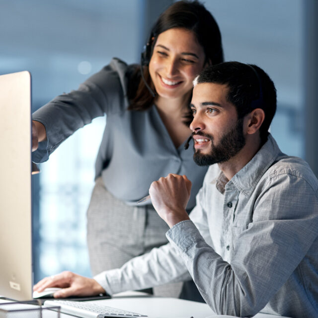 Two people work collaboratively at a desk, one standing and pointing at a computer screen while the other sits, using a keyboard and wearing headphones.