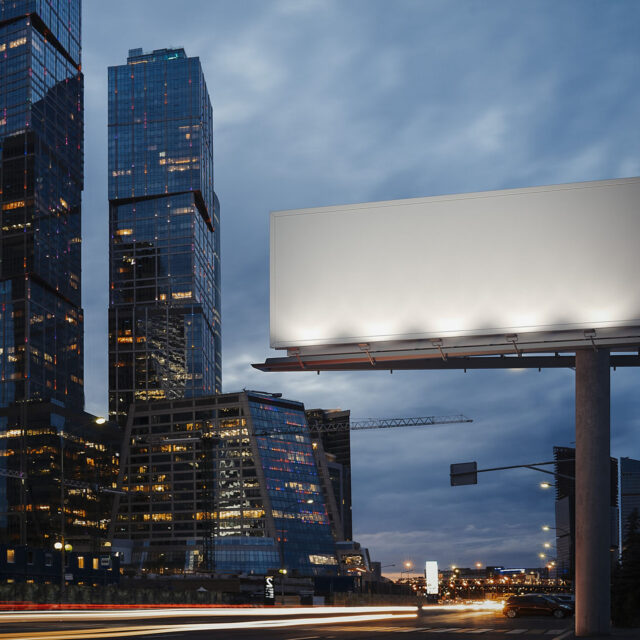 A blank, illuminated billboard stands in front of tall, modern buildings at dusk, with a busy street and light traffic below.