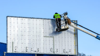 A worker in a high-visibility jacket is using an elevated platform to install or maintain an empty billboard against a clear blue sky.