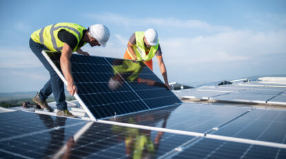 Two workers in safety gear install solar panels on a rooftop under a clear sky.