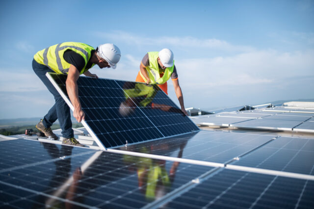 Two workers in safety gear install solar panels on a rooftop under a clear sky.