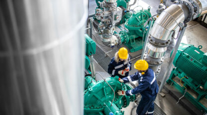 Two workers in protective gear and yellow helmets inspect large green machinery in an industrial facility. Stainless steel pipes and various mechanical components are visible in the background.