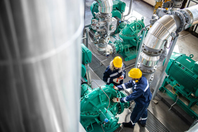 Two workers in protective gear and yellow helmets inspect large green machinery in an industrial facility. Stainless steel pipes and various mechanical components are visible in the background.