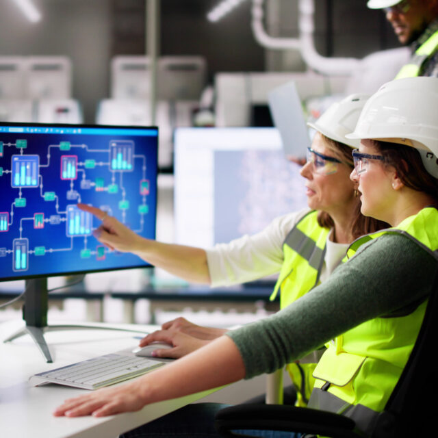 Two engineers wearing hard hats and safety vests work on a computer with a flowchart displayed on the screen.