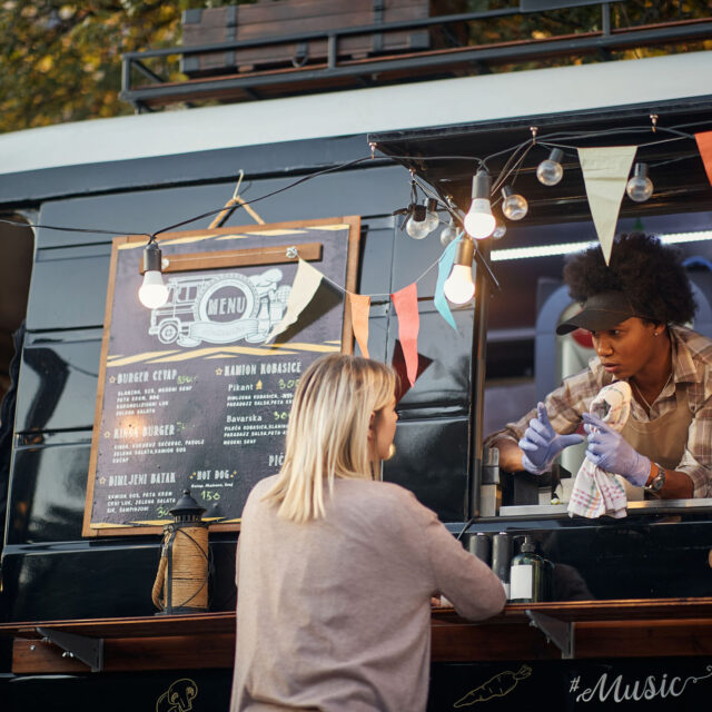 A woman stands at the counter of a black food truck, ordering food from a server. The truck has a chalkboard menu, hanging lights, and colorful flag decorations.