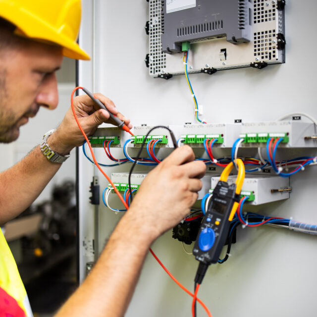A technician wearing a yellow hard hat and safety vest uses a multimeter to test electrical connections in a control panel.