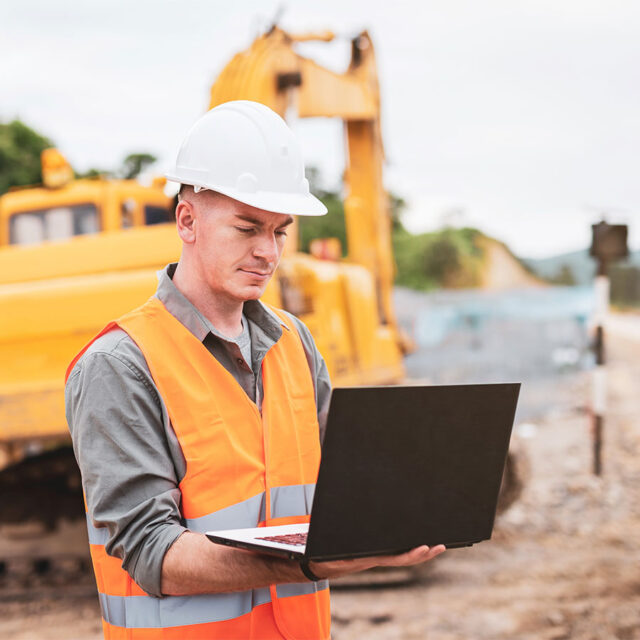 A construction worker in a white hard hat and orange safety vest uses a laptop at a construction site with heavy machinery in the background.