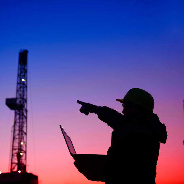 Silhouette of a worker wearing a hard hat, holding a laptop and pointing towards oil rigs at sunset.