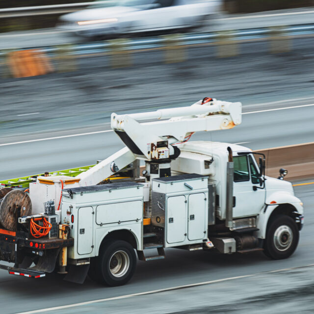 A white utility truck with an aerial lift and various equipment in the rear is traveling on a multi-lane road.