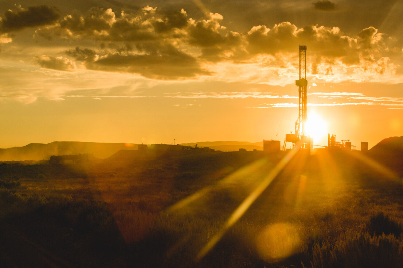 A drilling rig is silhouetted against a golden sunset in a remote landscape, with scattered clouds in the sky.