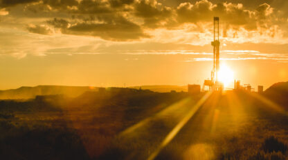 A drilling rig is silhouetted against a golden sunset in a remote landscape, with scattered clouds in the sky.
