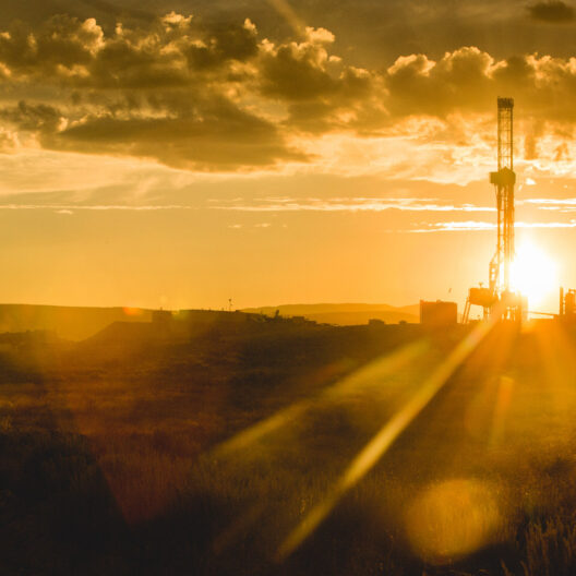A drilling rig is silhouetted against a golden sunset in a remote landscape, with scattered clouds in the sky.