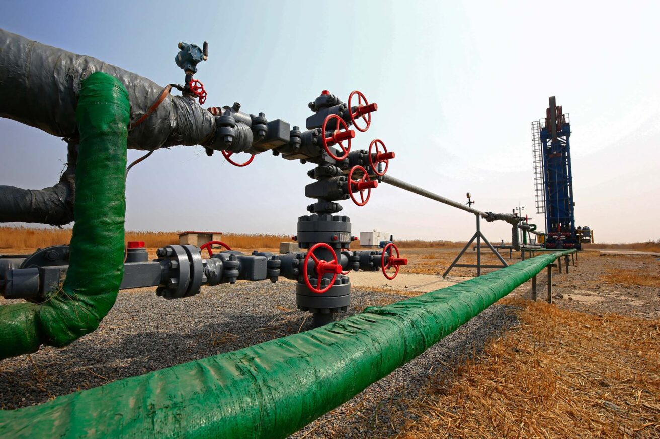 A natural gas pipeline system with multiple valves and a rig in the background stands in an open, dry landscape.
