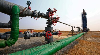 A natural gas pipeline system with multiple valves and a rig in the background stands in an open, dry landscape.