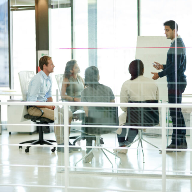 A man stands and speaks to four seated colleagues in a modern conference room with large windows.