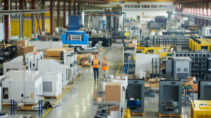 Two workers in orange safety vests and helmets walk through a large, organized industrial manufacturing facility with various machinery and equipment.