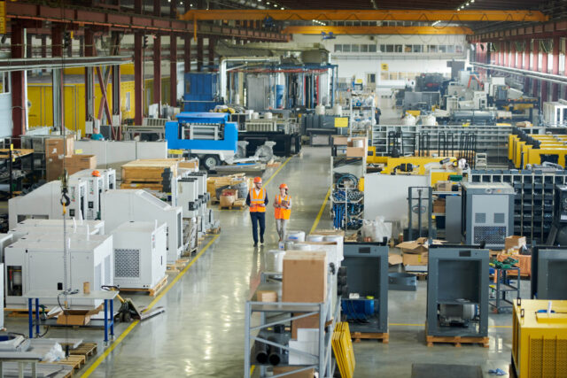 Two workers in orange safety vests and helmets walk through a large, organized industrial manufacturing facility with various machinery and equipment.