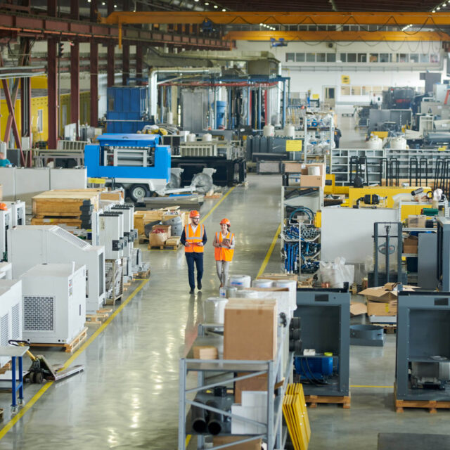 Two workers in orange safety vests and helmets walk through a large, organized industrial manufacturing facility with various machinery and equipment.