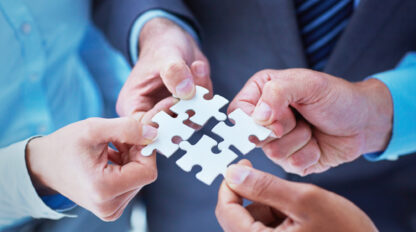 Four people in business attire are connecting four puzzle pieces together, symbolizing teamwork and collaboration.