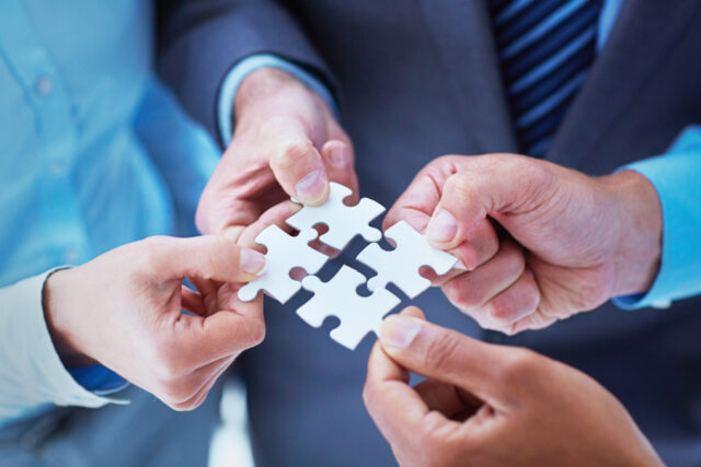 Four people in business attire are connecting four puzzle pieces together, symbolizing teamwork and collaboration.