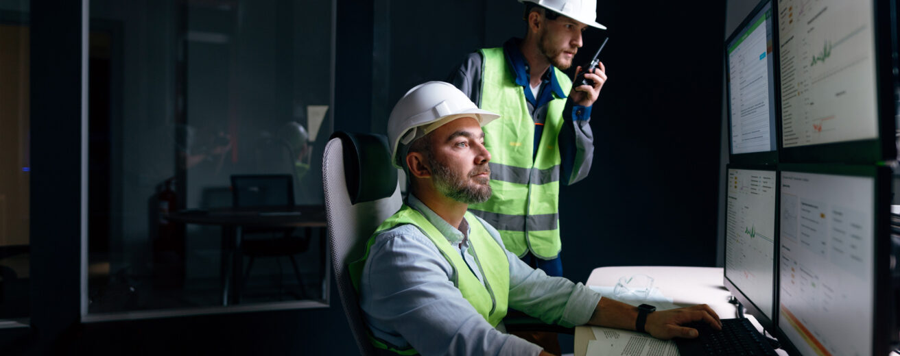 Two construction workers wearing safety vests and hard hats use computers; one is seated operating multiple screens, and the other stands behind using a walkie-talkie.