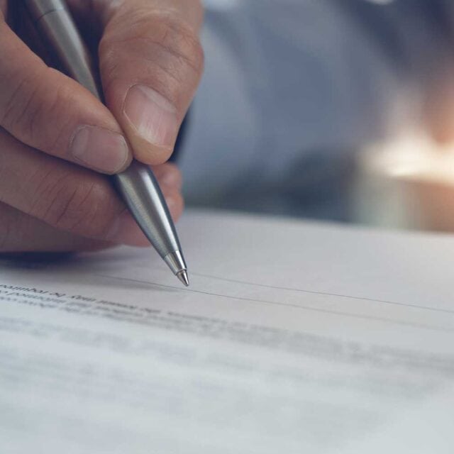 A person holding a pen is signing a document close-up. The image shows a hand with a pen hovering over a paper with printed text.