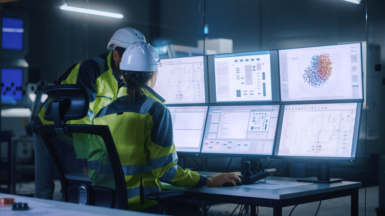 Two workers in safety gear monitor data and charts on multiple computer screens in a control room setting.
