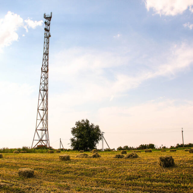 A rural landscape with telephone towers, bales of hay scattered on a grassy field, and a tree in the background under a partly cloudy sky.