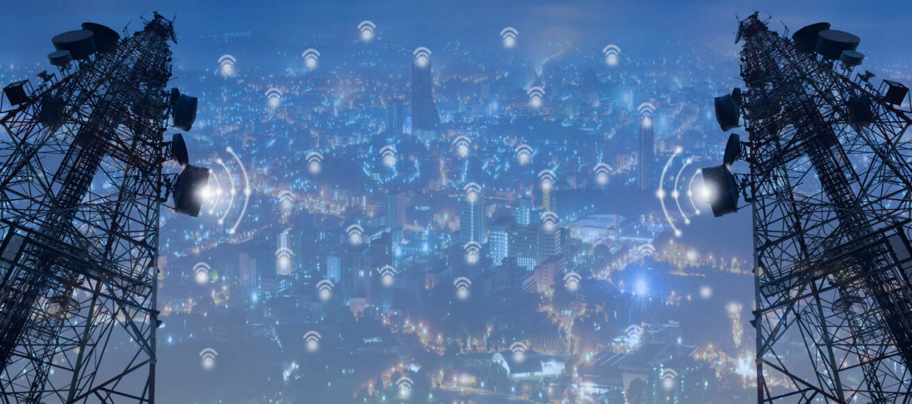 Two telecommunications towers stand on either side of an urban skyline filled with glowing lights, symbolizing widespread wireless network connectivity throughout the city.