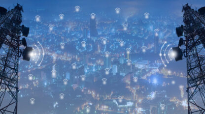 Two telecommunications towers stand on either side of an urban skyline filled with glowing lights, symbolizing widespread wireless network connectivity throughout the city.