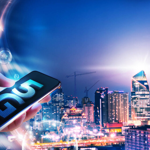 Hand holding smartphone displaying "5G" in foreground with a brightly lit, modern cityscape and communication beams in the background at dusk.