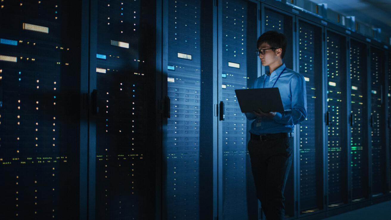 A person holding a laptop stands in a server room, monitoring the servers.
