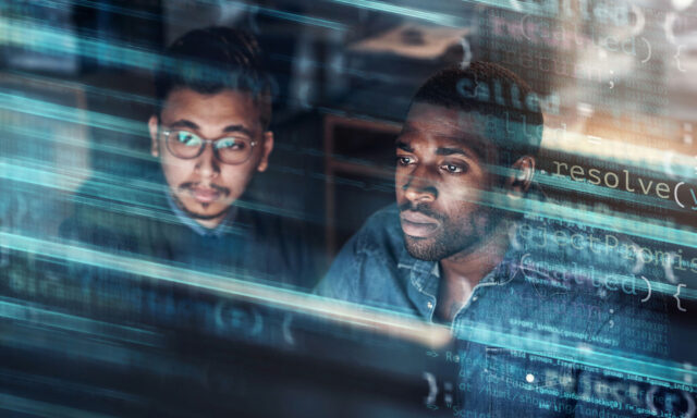 Two men focus on a computer screen filled with lines of code, suggesting they are engaged in software development or programming. .