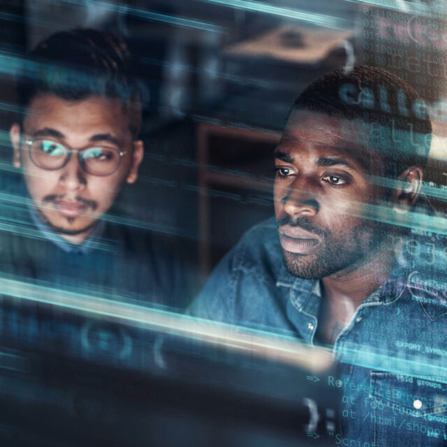 Two men focus on a computer screen filled with lines of code, suggesting they are engaged in software development or programming. .