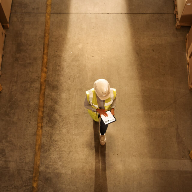Top-Down View: Worker Wearing Hard Hat Checks Stock and Inventory Using Digital Tablet Computer in the Retail Warehouse full of Shelves with Goods. Working in Logistics, Distribution