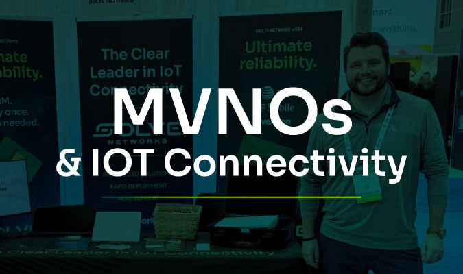 Man smiling at a tech booth with banners reading "MVNOs & IoT Connectivity" and "The Clear Leader in IoT Connectivity."